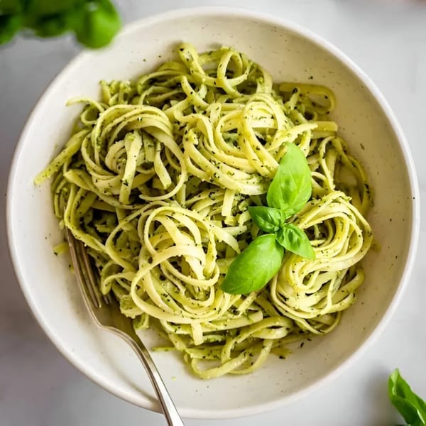 A white bowl filled with green pesto pasta, garnished with fresh basil leaves and a fork.