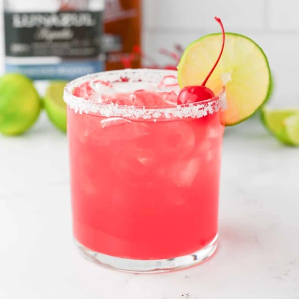 A vibrant red cocktail with a salted rim, garnished with a lime slice and a cherry.