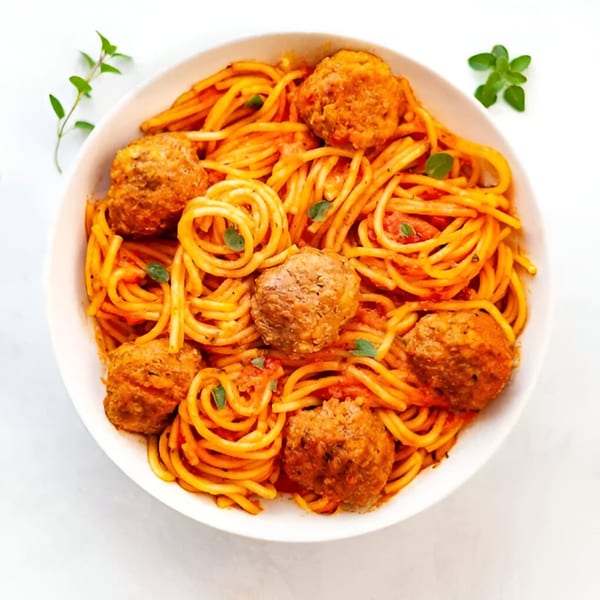 Spaghetti with meatballs and tomato sauce, garnished with fresh herbs in a white bowl.