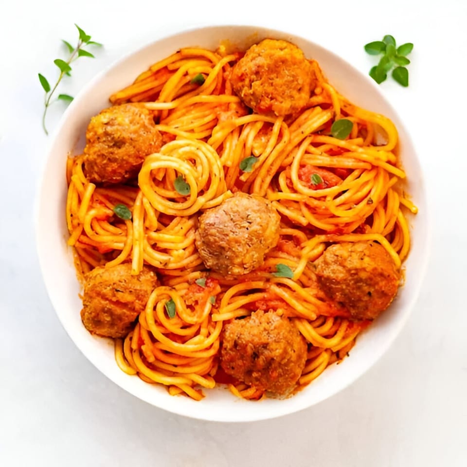 Spaghetti with meatballs and tomato sauce, garnished with fresh herbs in a white bowl.